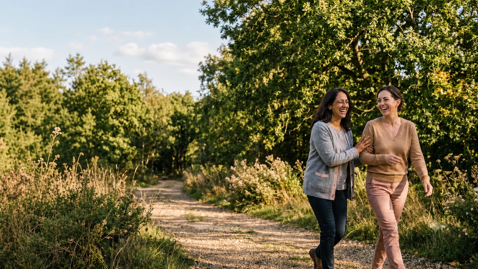 Two women laughing together on a sunlit, tree-lined path
